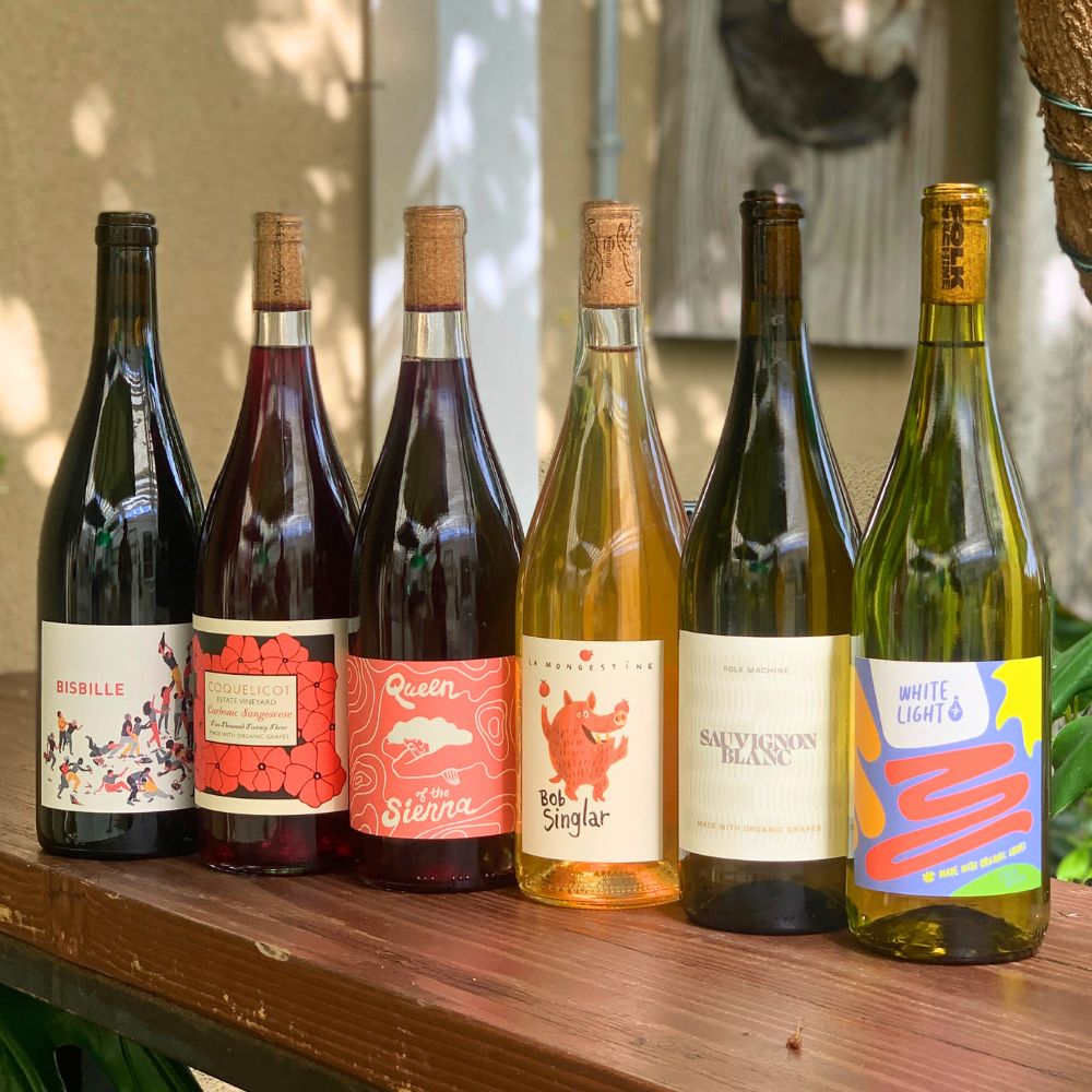 Row of natural wine bottles with colorful labels on a wooden surface outdoors.